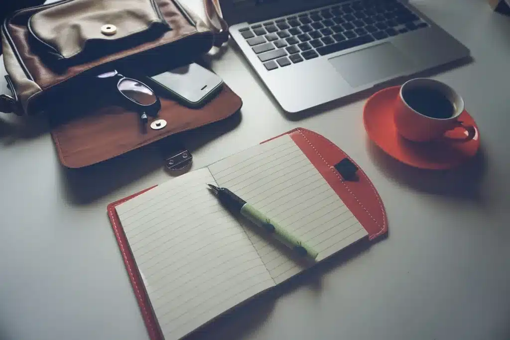 Mains tenant un carnet de voyage et une carte posés sur une table de café, sac et ordinateur flous en arrière-plan